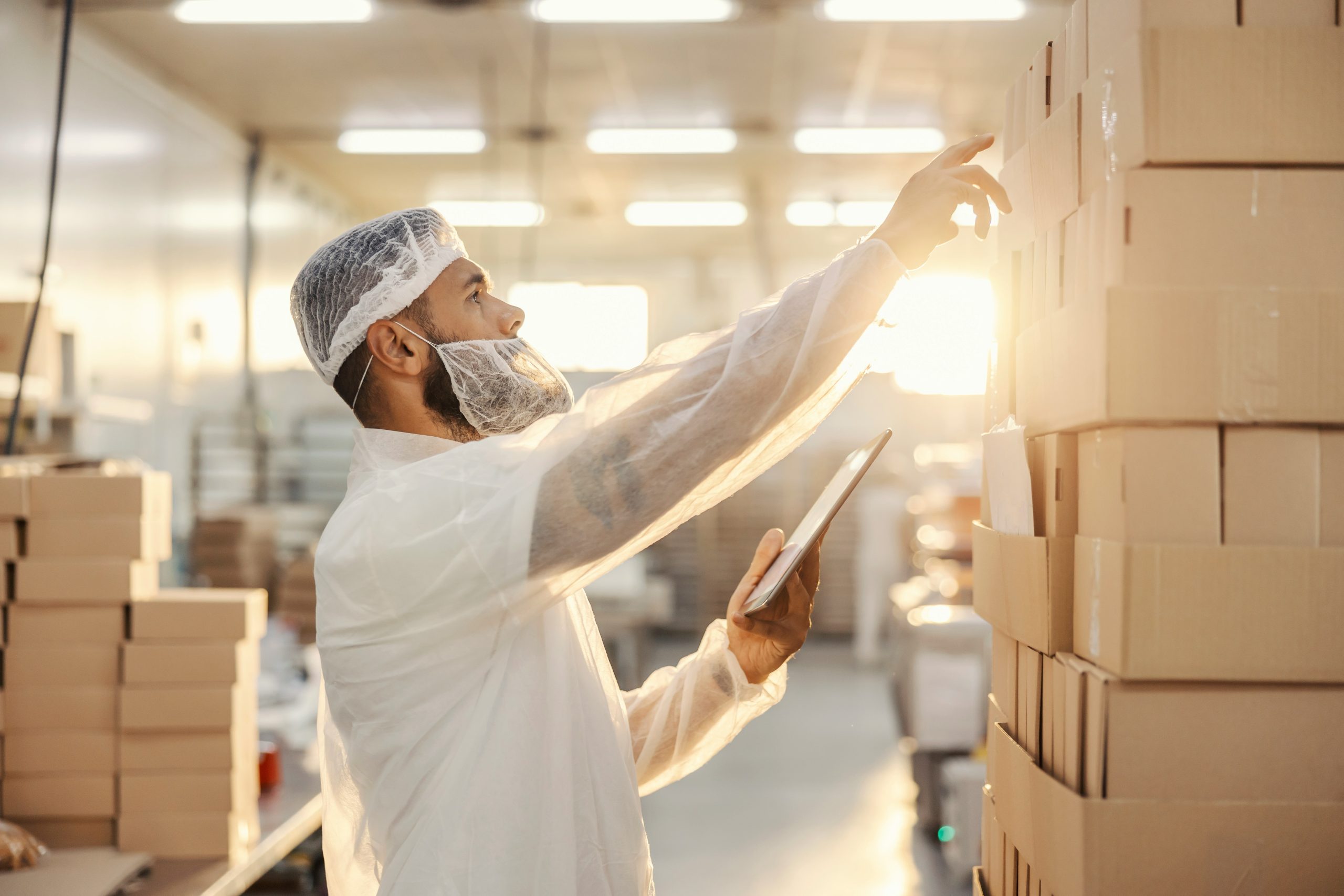 A worker dressed in sanitary protective gear reviews inventory by inspecting stacked cardboard boxes and entering data on a tablet inside a food manufacturing facility, illuminated by warm sunlight.