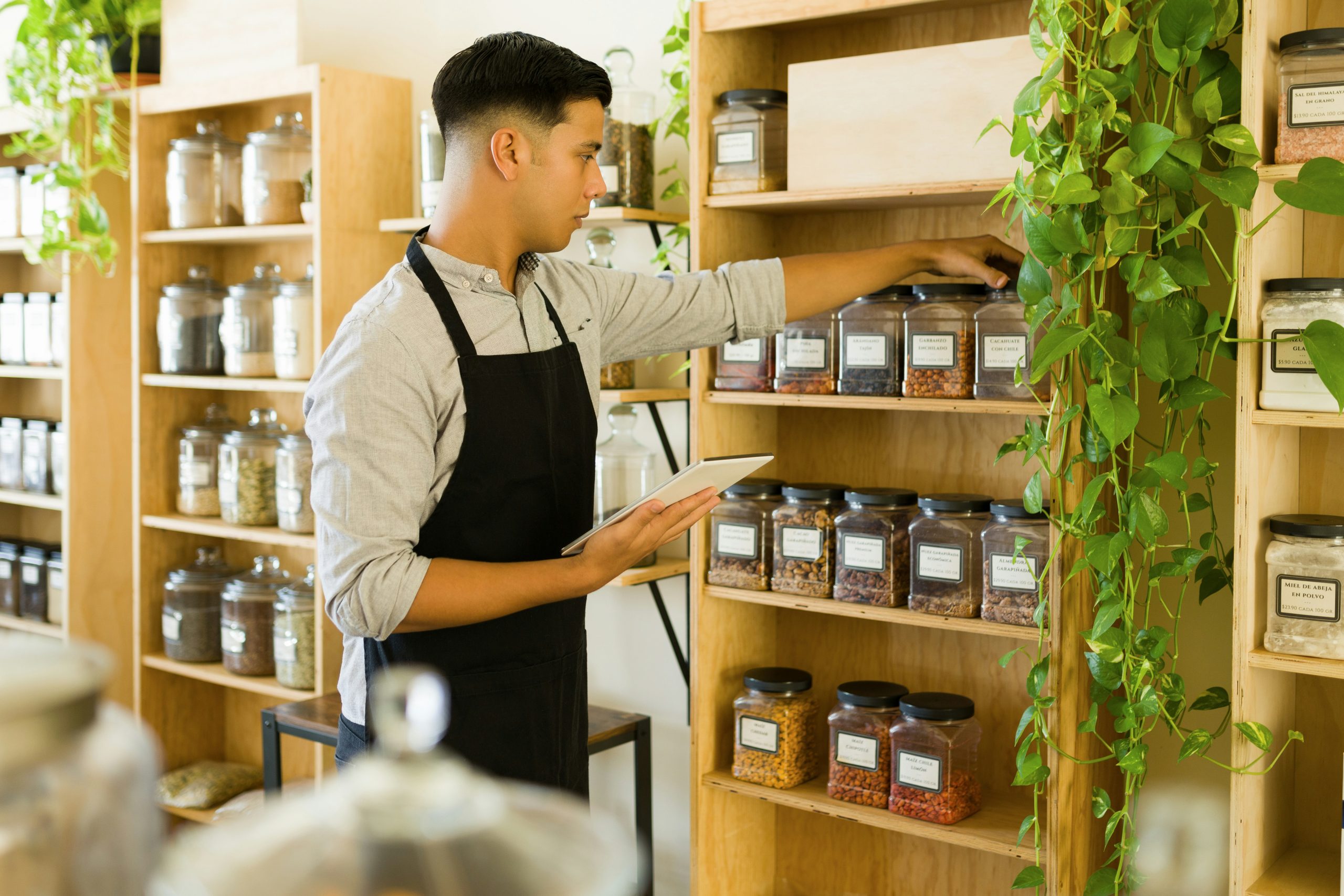 A store associate uses a tablet to check and organize inventory across shelves of labeled jars—illustrating the manual, time-consuming stock processes that Nvexus helps automate and streamline.