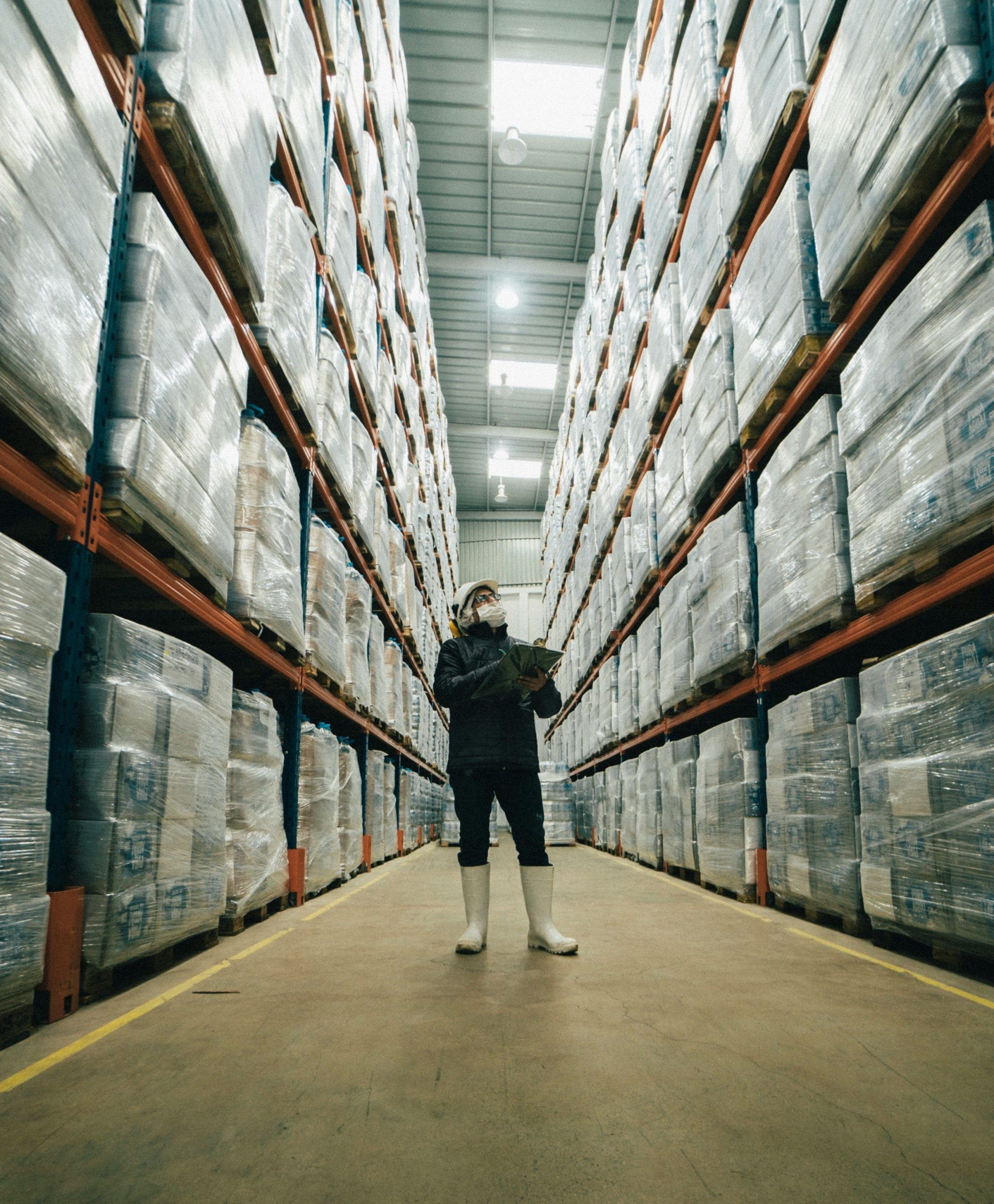 A warehouse employee performs a manual inventory check in a tall pallet rack aisle—illustrating the visibility gaps, cycle-count challenges, and on-hand accuracy issues that Nvexus helps automate for distribution and manufacturing teams.