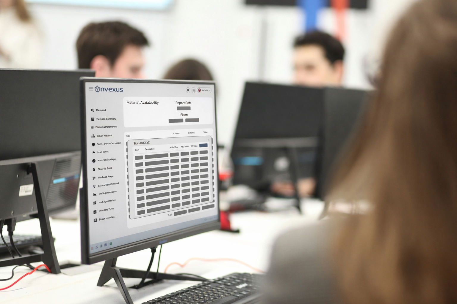 Office workstation displaying the Nvexus Material Availability module, showing item-level availability, ABC classifications, and supply status while colleagues collaborate in the background.
